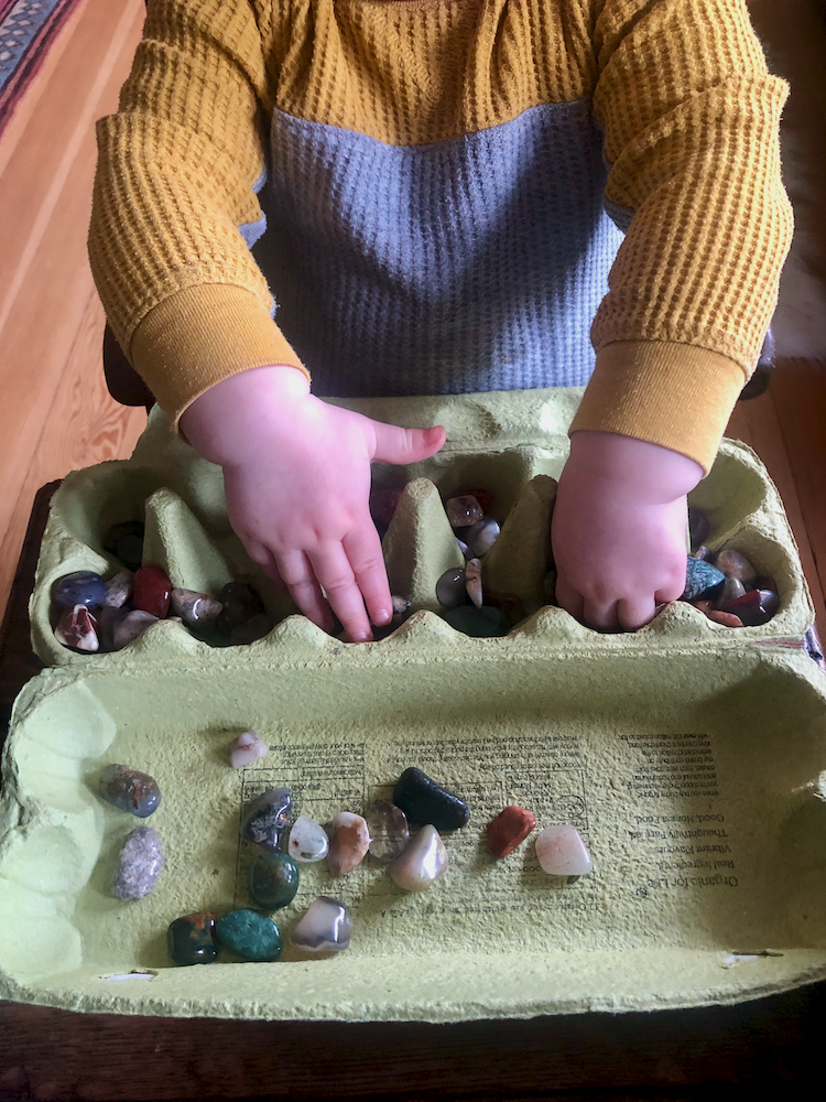 Toddler exploring an egg carton filled with healing crystals, touching and choosing stones with both hands.
